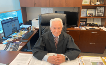 Judge Jim Booras sits at his desk. He is an older man with white hair. He is smiling. His desk is covered with periodicals and memos.