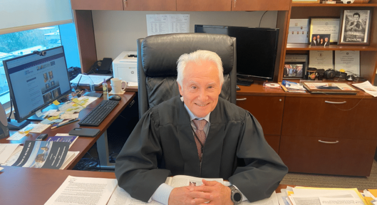 Judge Jim Booras sits at his desk. He is an older man with white hair. He is smiling. His desk is covered with periodicals and memos.